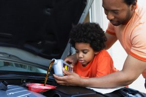 Um homem adulto vestindo uma camisa na cor laranja claro e uma criança vestindo uma camisa na cor laranja escuro colocando juntos o óleo no motor.