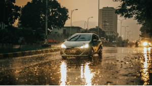 Carro cinza em movimento sob a chuva, com faróis acesos e gotas d'água visíveis na lataria, cruzando uma pista molhada e refletindo no asfalto.