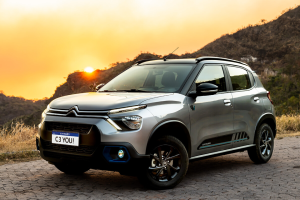Citroën C3 cinza estacionado em ambiente arborizado, com árvores ao fundo e céu alaranjado de pôr do sol iluminando a cena.