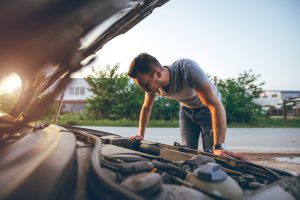 Homem verificando o motor de um carro com o capô aberto, em ambiente externo, observando os componentes do veículo.