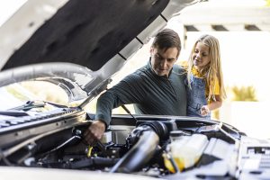 Homem com uma menina no colo observando a correia dentada do carro com o capô aberto.