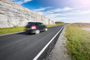 Carro preto em movimento na estrada. Em destaque, céu azul ensolarado com nuvens e grama dos dois lados da estrada. Ao lado esquerdo, um muro grande de pedras naturais na cor cinza.