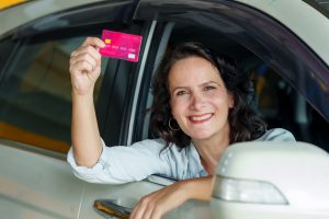 Caucasian woman adult leaning out of car window smiling holding up credit card. light denim jacket. Indicates satisfaction after completing a car service or insurance transaction at a repair shop.