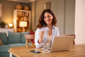 Happy mid-adult woman holding a credit card and making an online purchase using her laptop at home, smiling brightly while looking at the card with joy and satisfaction
