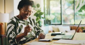 Happy, black woman and finance with laptop for payroll, tax return or budget planning at home. Female person, accounting or smile with calculator, documents or bills for expenses, deductions or audit