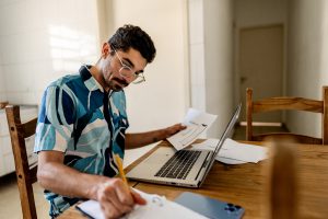 Focused man works on laptop at residential kitchen table surrounded by documents and papers, showing contemporary remote work model, professional flexibility, productivity in domestic environment, balance between personal life and career and adaptation to new distance work format