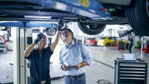 Female Mechanic And Manager Engage In Discussing Repairs Using Light Underneath A Car At Auto Repair Shop