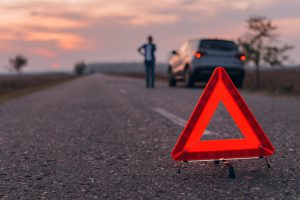 Warning triangle sign on the road, woman in blur background calling for roadside assistance by the broken car, selective focus