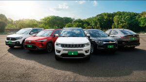 Cinco SUVs enfileirados e centralizados, da esquerda para a direita nas cores branca, vermelha, branca, preta e cinza. Ao fundo, árvores e céu azul ensolarado.