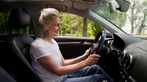 Mulher madura no volante, em um ambiente arborizado e aberto.