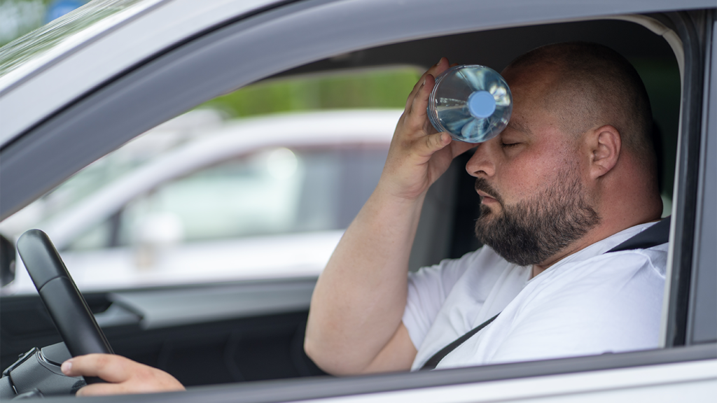 Motorista é visto se refrescando com garrafinha de água, com carro parado em estacionamento. 