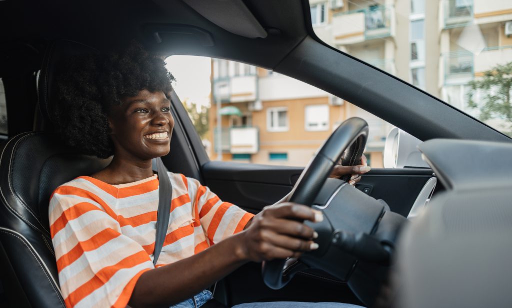 Mulher negra, sorridente, vestindo uma blusa com listras brancas e laranjas, usando cinto de segurança, dirigindo um carro.