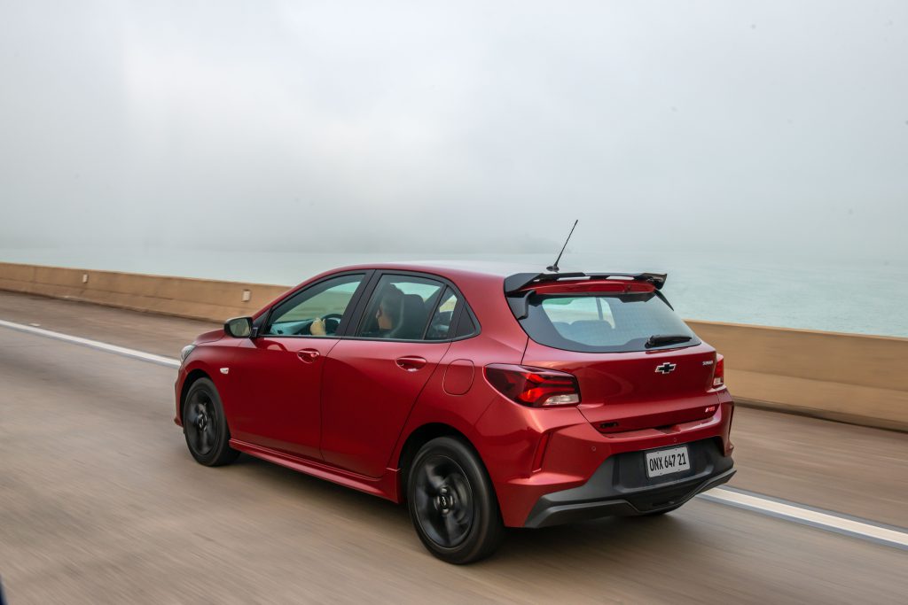 Traseira do Chevrolet Onix vermelho em movimento, com lanternas acesas e design aerodinâmico em evidência, em cenário de estrada