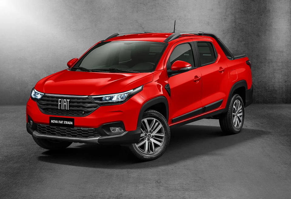Nova Fiat Strada vermelha vista de frente e lateral, com design robusto, faróis em LED e grade frontal renovada, estacionada em ambiente urbano.