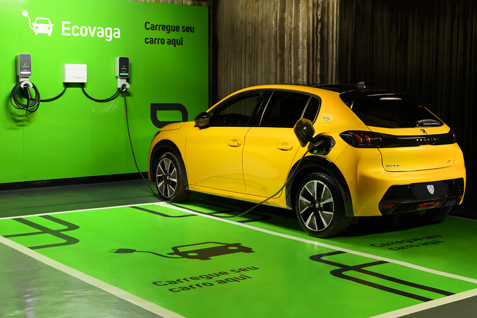 Peugeot 208 amarelo elétrico sendo carregado em estação, com cabo conectado na lateral do carro.