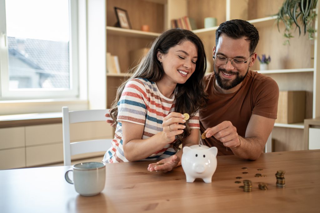 Casal sorridente colocando moedas em um cofrinho de porquinho, simbolizando economia e planejamento financeiro.