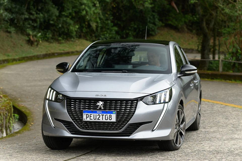 Peugeot 208 cinza em movimento na cidade, com um homem ao volante visível pelo vidro.