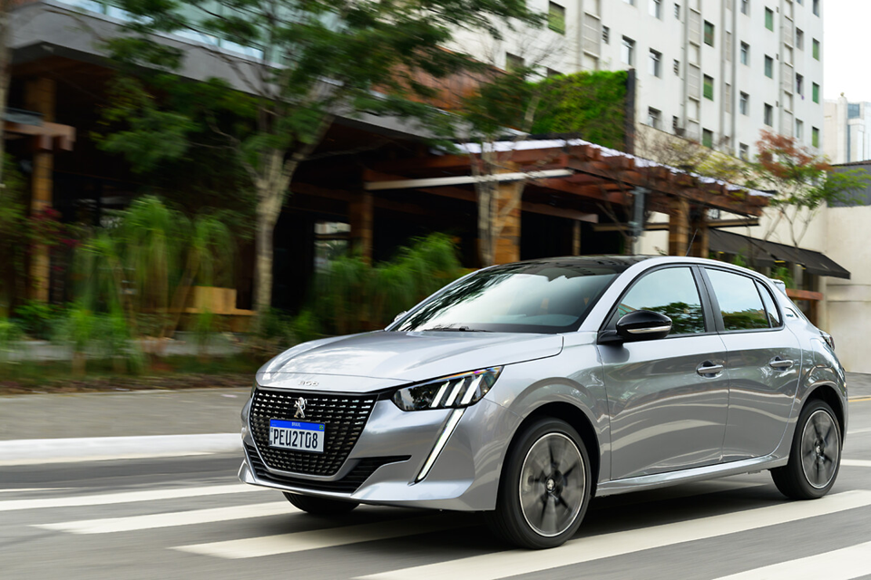 Peugeot 208 cinza em movimento na cidade, com rodas girando e design moderno em destaque.