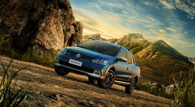 "Volkswagen Saveiro azul estacionada em ambiente rural, cercada por árvores e montanhas ao fundo.