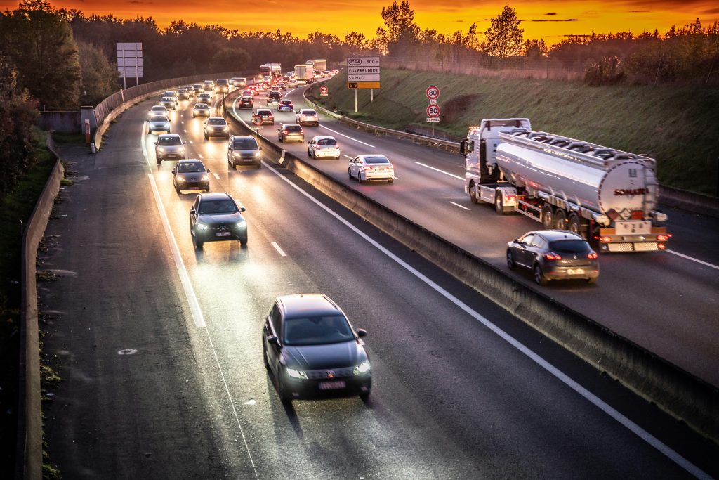 Estrada com carros em mão dupla. Ao fundo e nas laterais, montanhas e por do sol em destaque.
