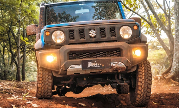 Suzuki Jimny azul em trilha de floresta, levantando poeira, cercado por árvores e vegetação.