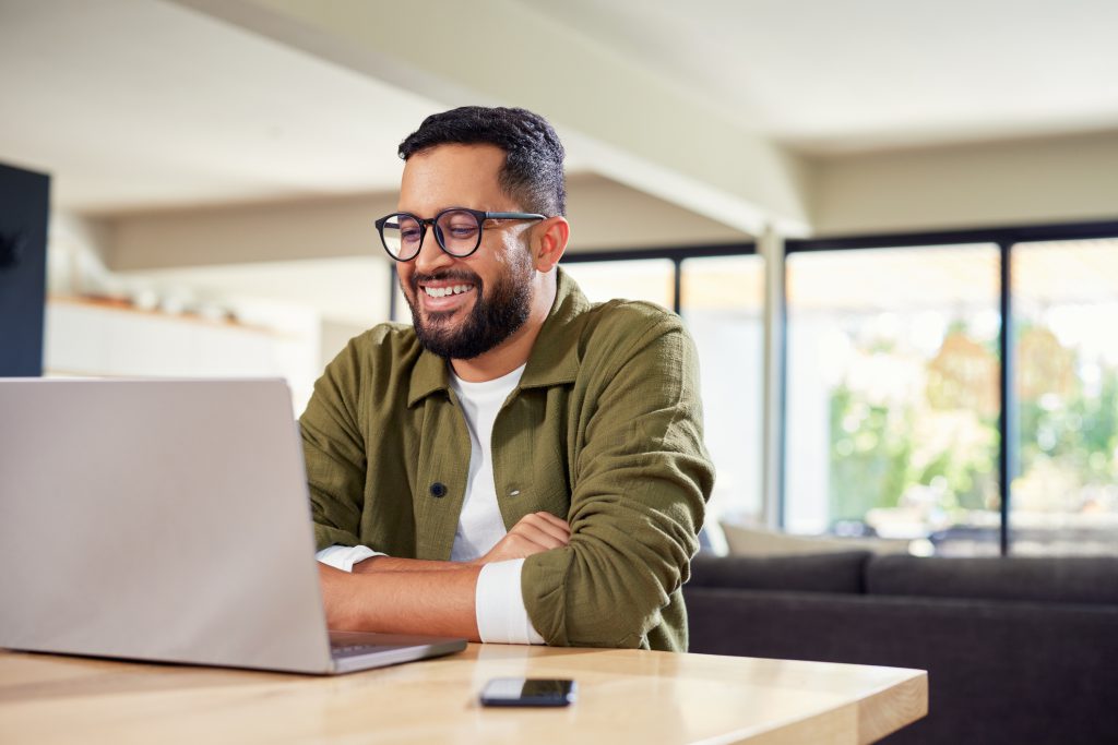 Smiling young Indian man sitting at home while using laptop. Multiethnic business man attending meeting online at home while using laptop. Happy mixed race man wearing eyeglasses while using laptop during work from home with copy space.