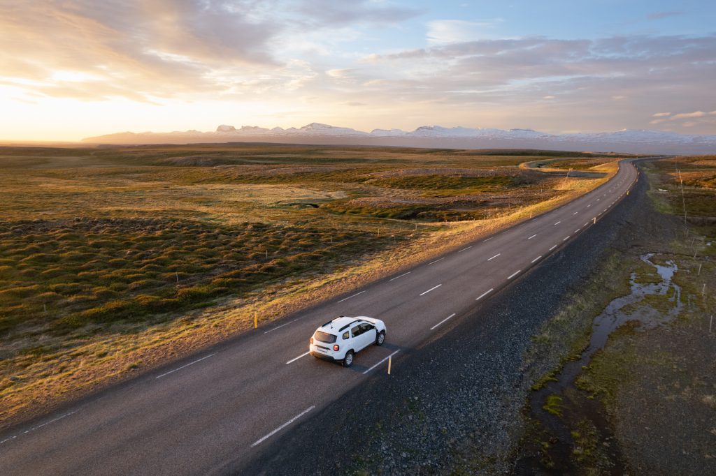 A vehicle on a winding road along a beautiful sunset surrounded by nature and mountains in distance. Aerial view of serene and peaceful scenery depicting travel, exploration, and tranquil rural life