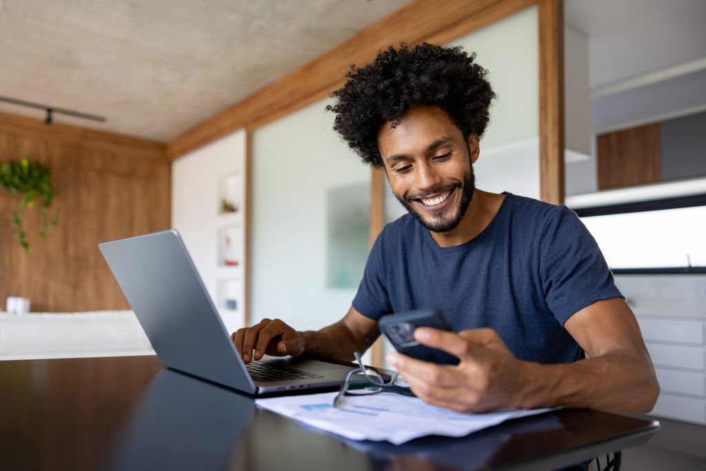 Happy Brazilian entrepreneur working at home using his laptop and his cell phone and smiling - home office concepts