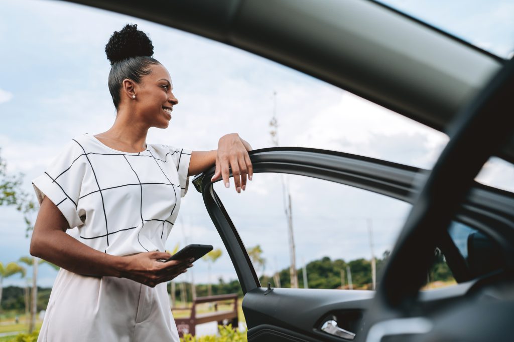 mulher com a porta do carro aberta segura o celular na mão, expressão de satisfação ao lado de um carro moderno