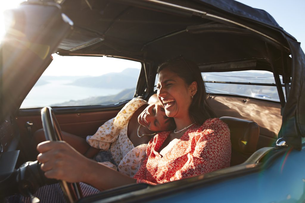 Duas mulheres felizes dentro de um carro em movimento, cabelos ao vento, sorrindo enquanto dirigem em uma estrada ensolarada