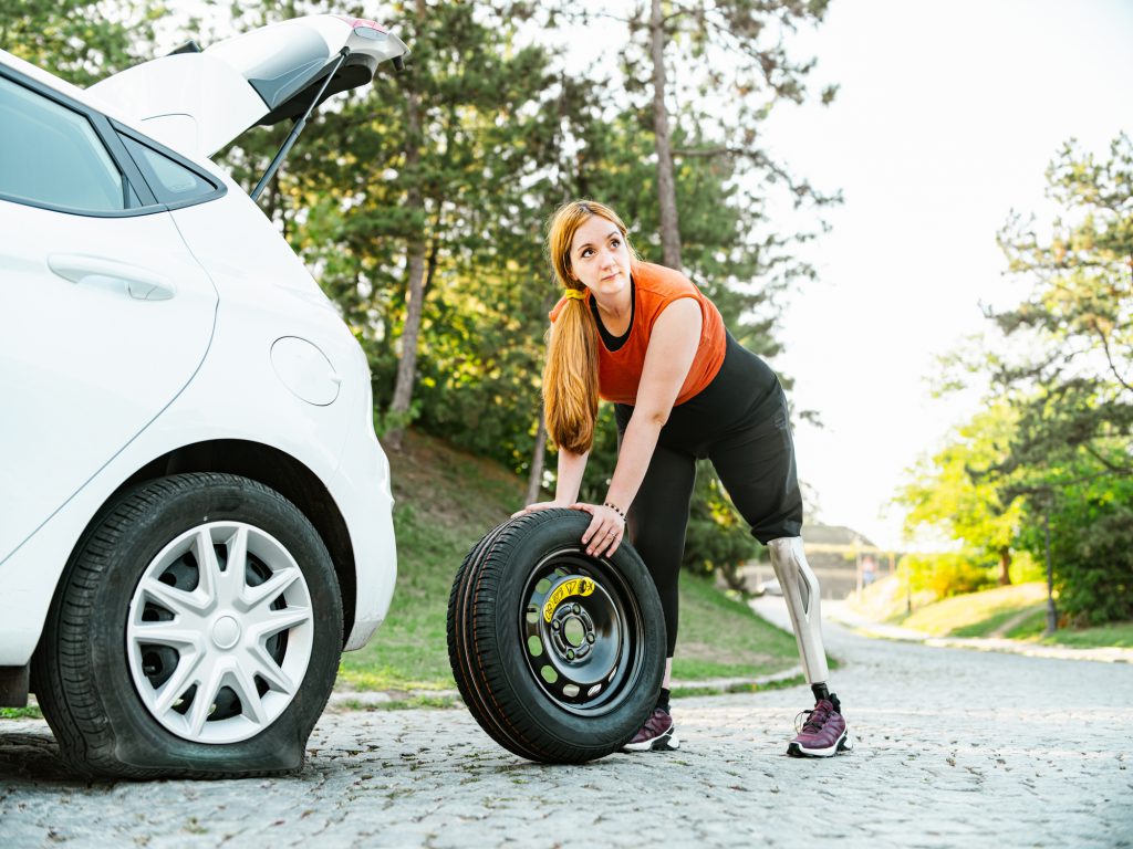 Young Woman, Leg Amputee having flat tire on the car. Young Caucasian woman, dressed in casual summer clothes with long red hair. Exterior of countryside, small white car on the side of the road.