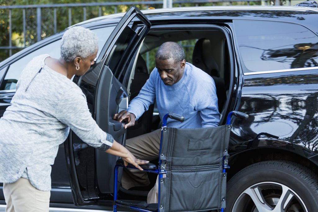 A disabled senior African American man is getting out of a car, into his wheelchair.  His wife is helping him, holding the door open.  Focus on man.