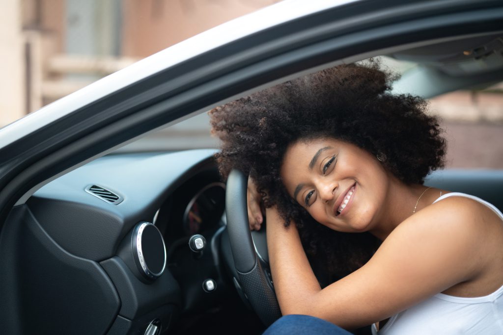 Mulher sentada no carro, sorrindo e abraçada ao volante, transmitindo sensação de segurança e satisfação ao dirigir.
