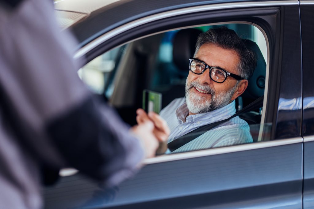 A senior man with glasses sits in a car, smiling as he shows his ID to an officer. The scene conveys a sense of trust and authority interaction.