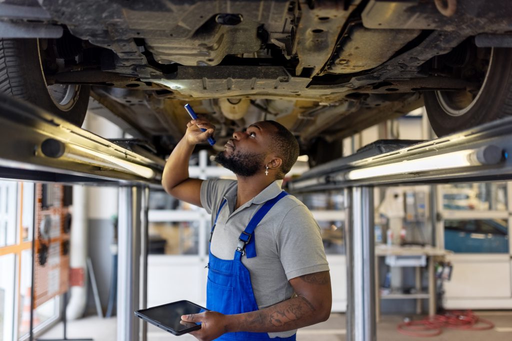 Repairman using torch while checking car from below at service station. Auto repair specialist examining brake pads of lifted car during inspection.